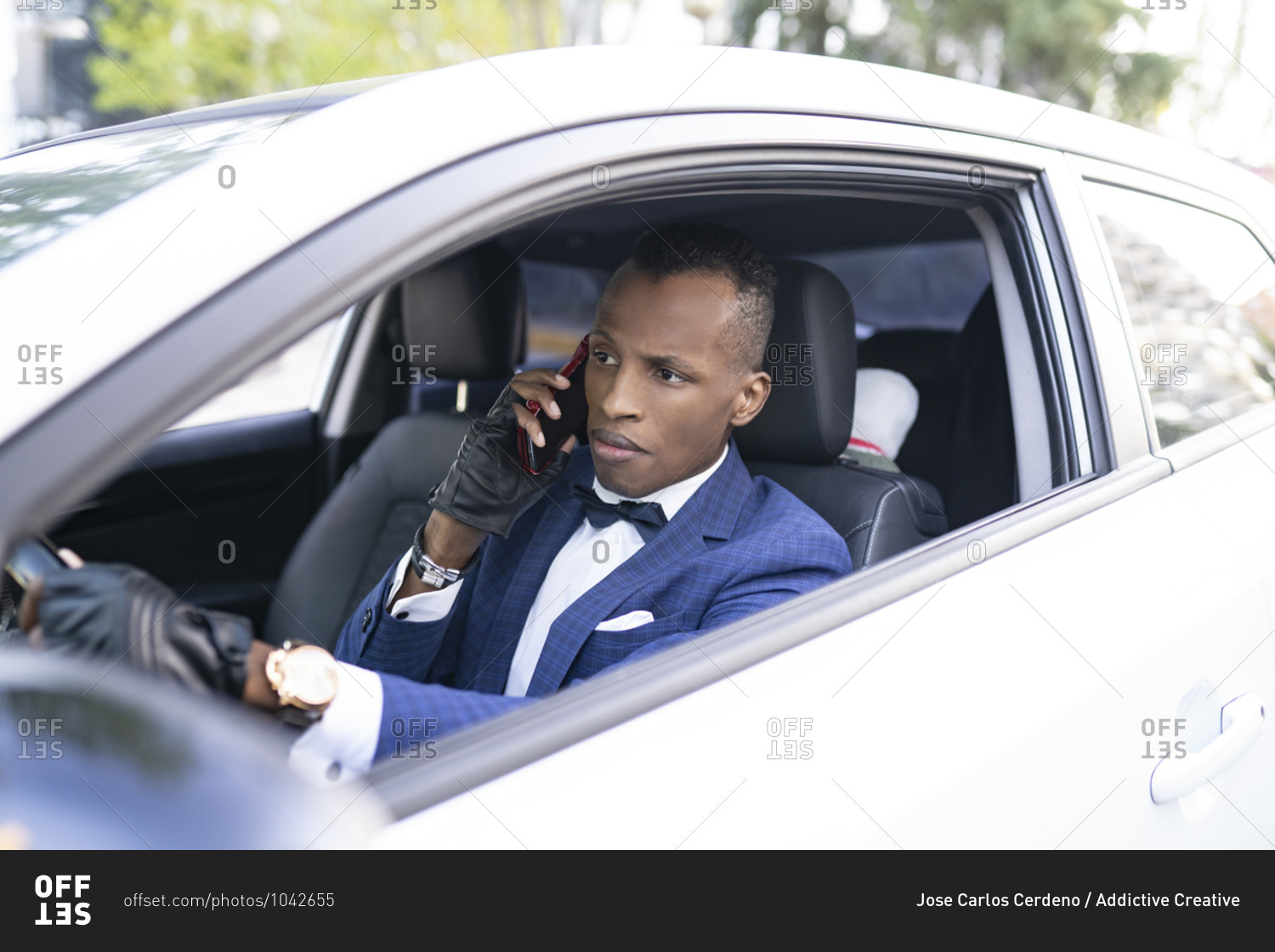 Serious ethnic male entrepreneur in classy suit with bow tie driving luxury car while talking on smartphone and looking away