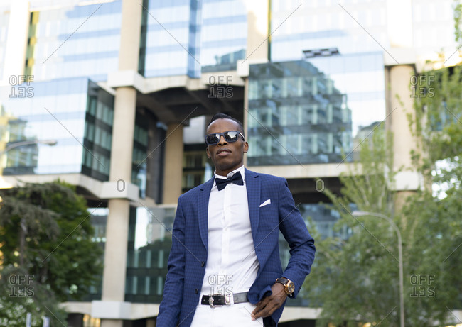 Low angle of masculine African American male entrepreneur wearing elegant suit and sunglasses standing in city and looking away