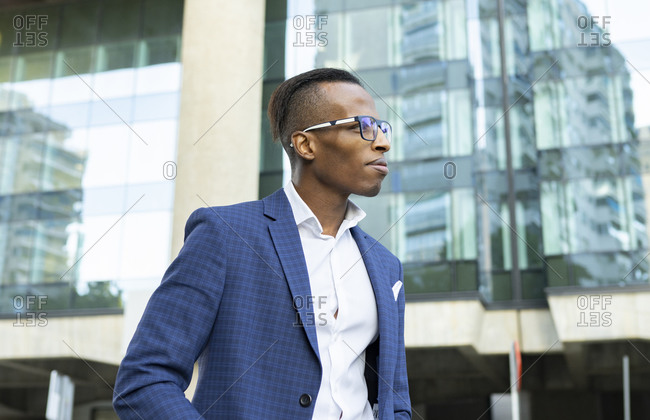 Low angle of masculine African American male entrepreneur wearing elegant suit and sunglasses standing in city and looking away