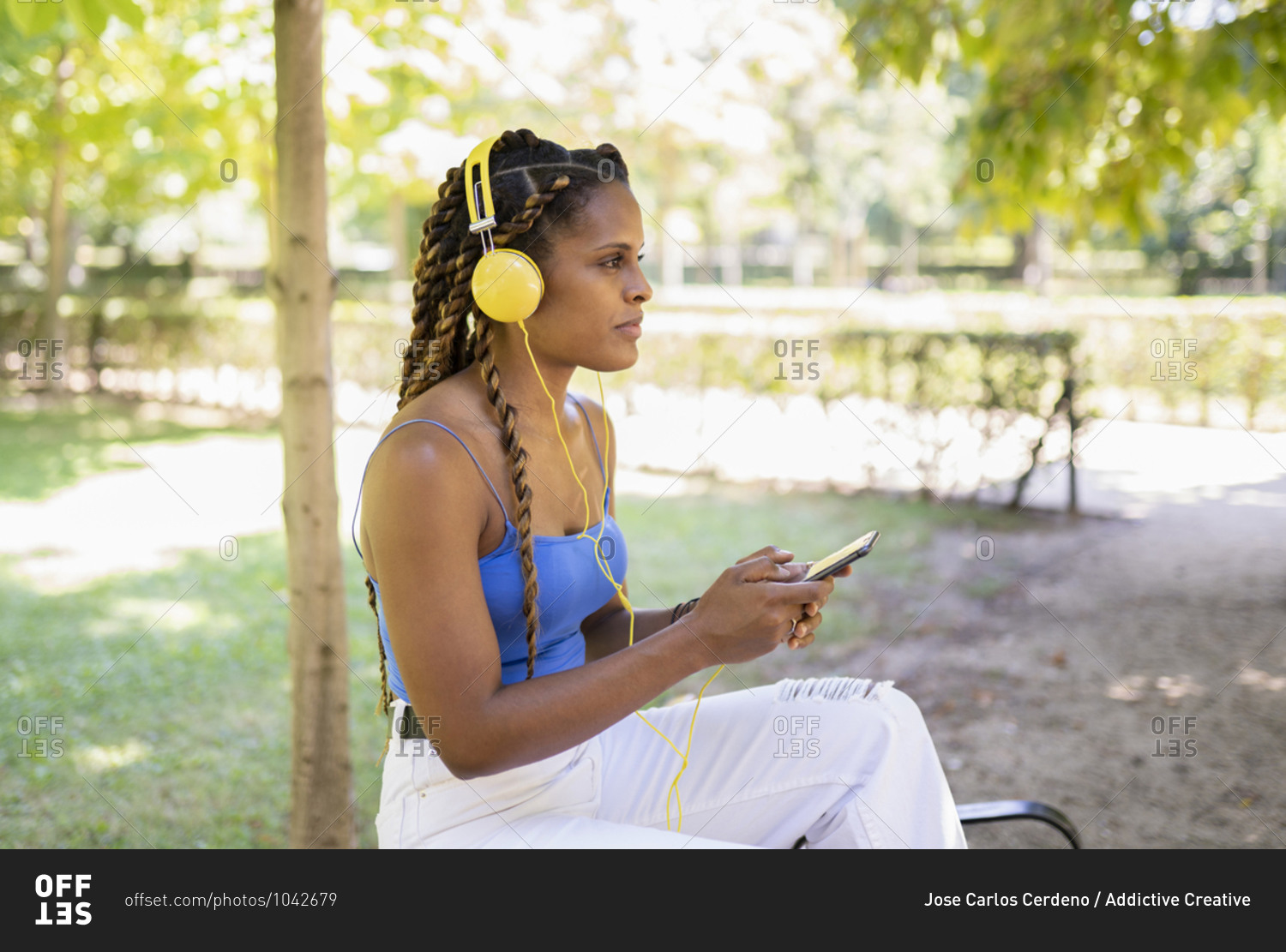 Positive Hispanic female listening to music in headphones while standing in urban park on sunny day and looking away