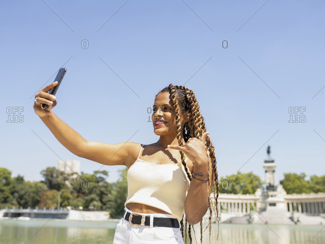 Smiling Hispanic female with trendy hairstyle with braids standing in city and taking photo on selfie camera of smartphone at weekend on sunny day