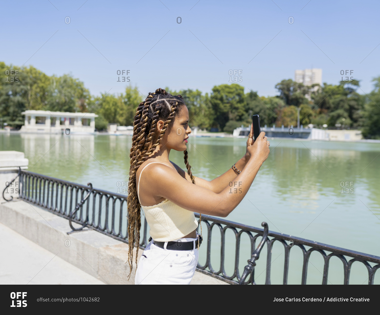 Side view of young ethnic female in summer wear and with braids standing near urban lake and taking picture on smartphone of amazing scenery in park