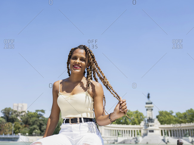 Carefree Hispanic female in trendy outfit and with braids sitting fence near lake in urban park and looking at camera