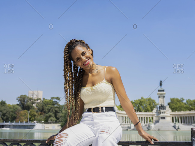 Carefree Hispanic female in trendy outfit and with braids sitting fence near lake in urban park and looking at camera