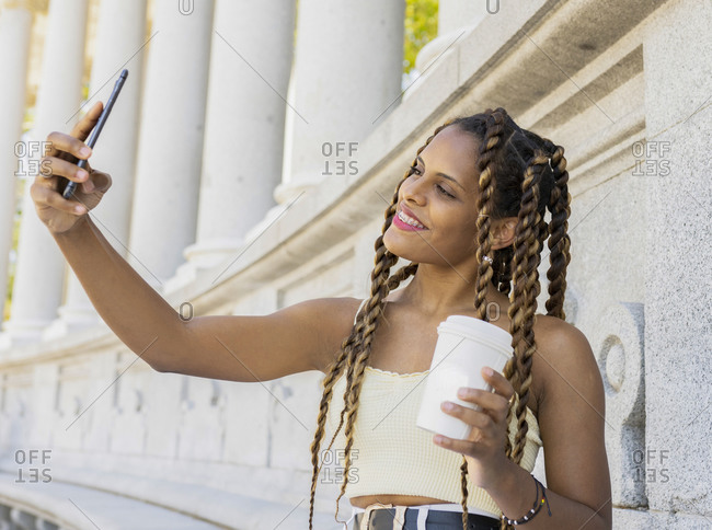 Carefree Hispanic female with braids and takeaway coffee taking self portrait on smartphone while enjoying city stroll in summer