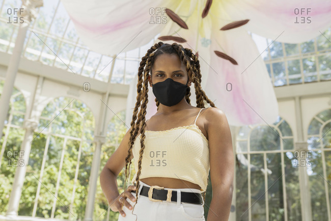 Trendy ethnic female with braids wearing protective mask standing in glass building lit by sunlight while looking at camera
