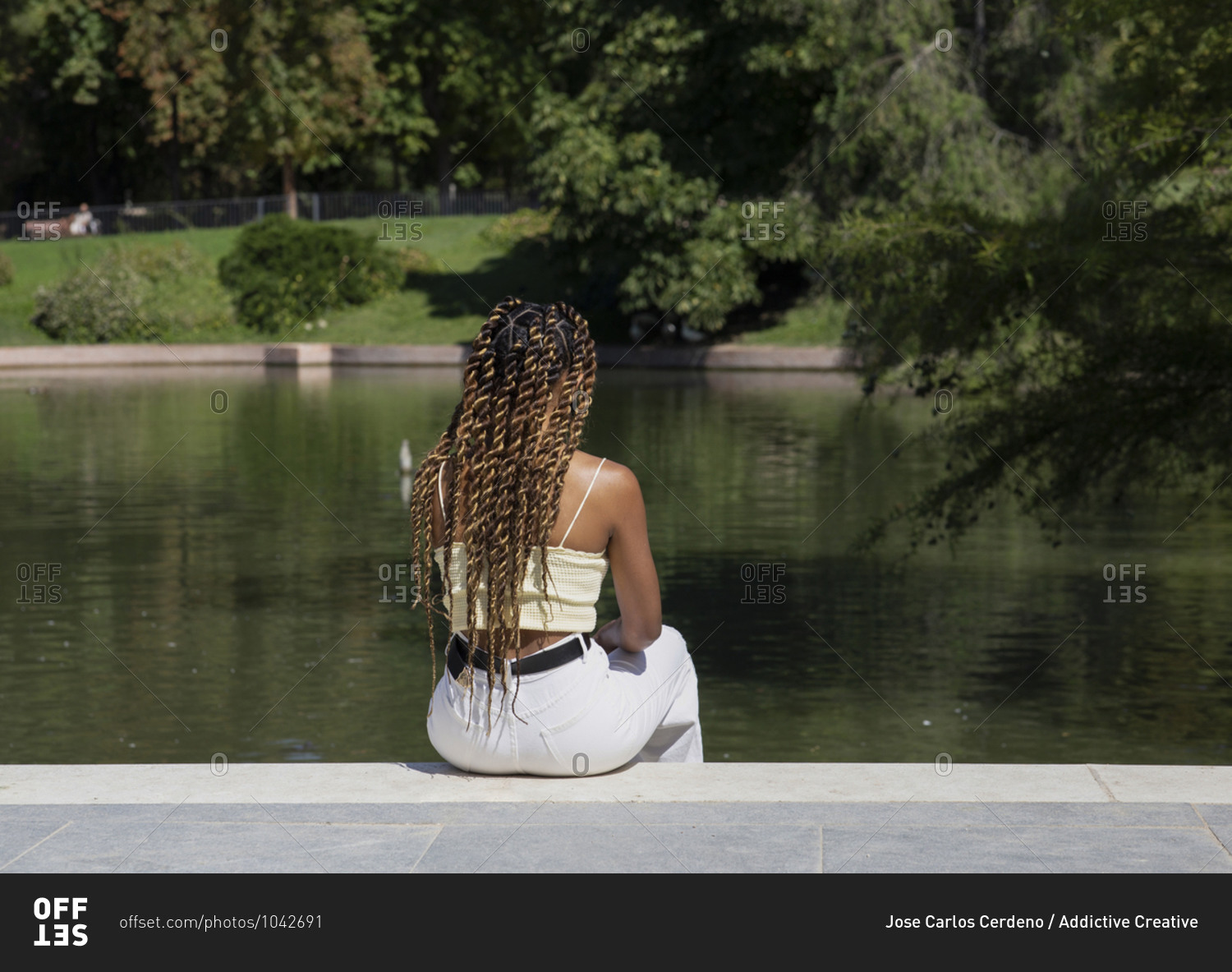 Back view of unrecognizable stylish ethnic female relaxing on embankment near pond and spending weekend in urban park in summer