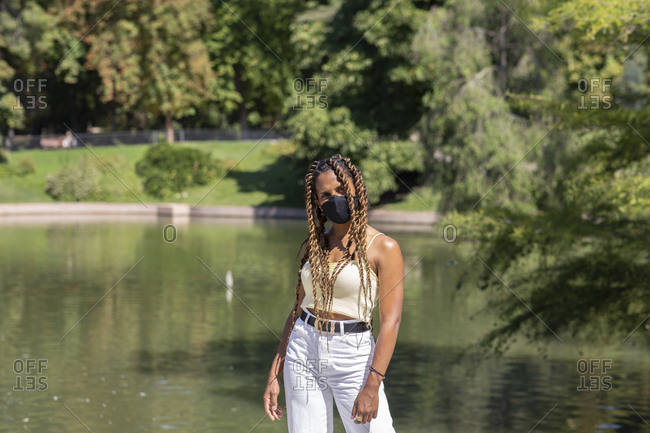Stylish ethnic female wearing medical mask during coronavirus standing near lake in park and reading messages on smartphone during stroll at weekend