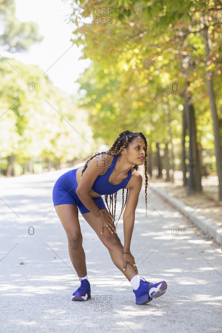 Confident Hispanic female athlete in sportswear doing forward bend and stretching legs during training in park while looking away