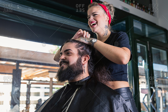 Side view of professional hipster tattooed female master setting long hair of stylish bearded man while working in modern barbershop