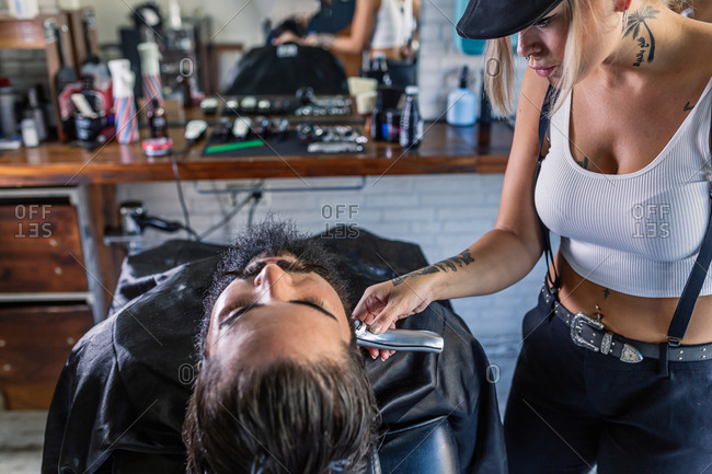 Side view of stylish tattooed female master shaving bearded customer with electric machine while working in professional barbershop
