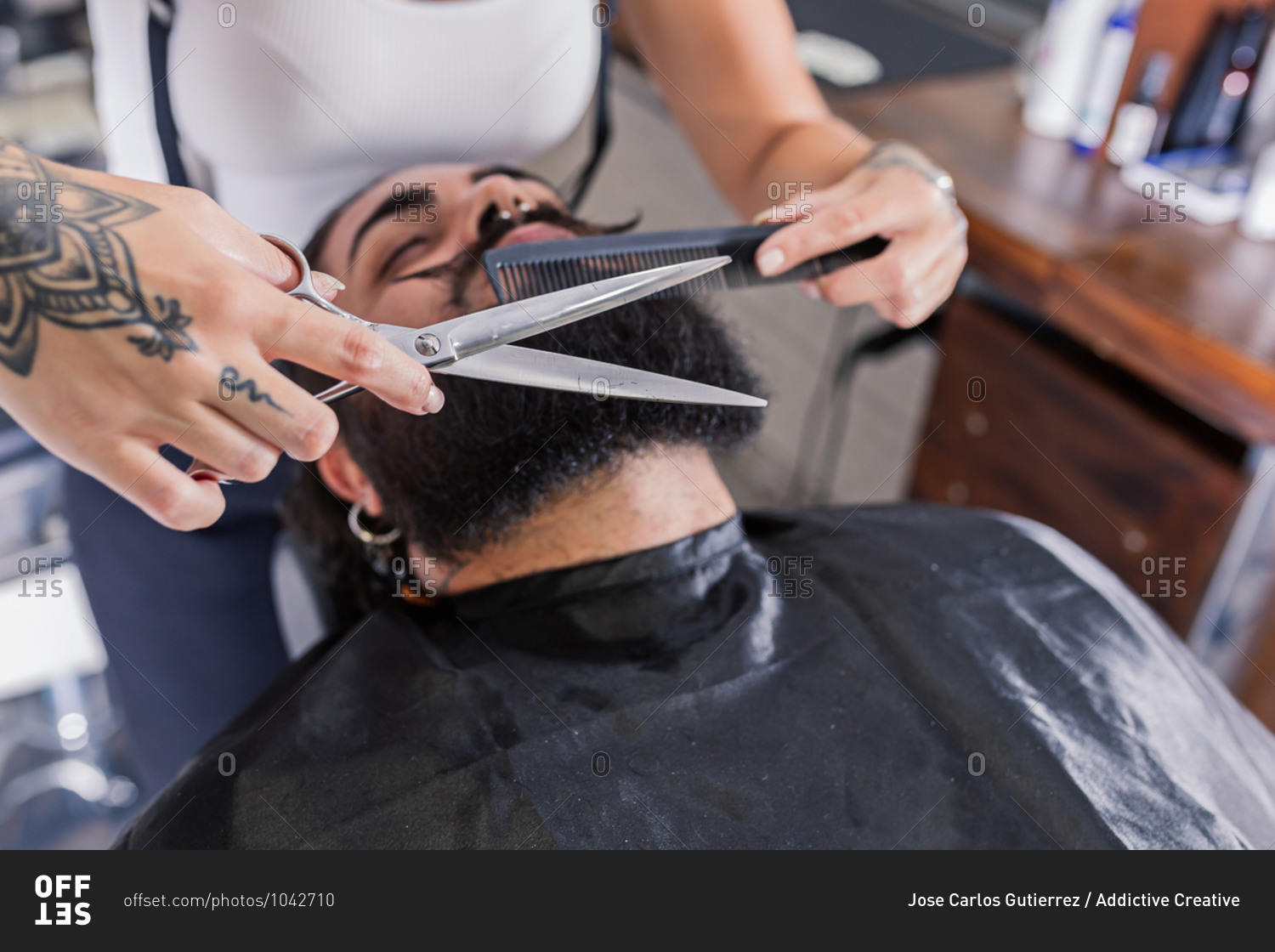 High angle of crop unrecognizable tattooed barber with scissors and comb cutting beard of stylish man sitting in chair in modern salon