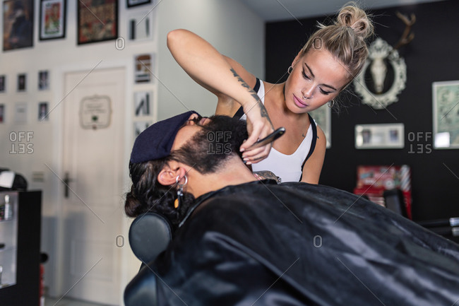 Side view of stylish tattooed female master shaving bearded customer with razor while working in professional barbershop