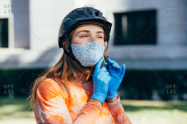 Female cyclist in latex gloves and fabric mask looking away and fastening hardhat on blurred background of street of Madrid, Spain