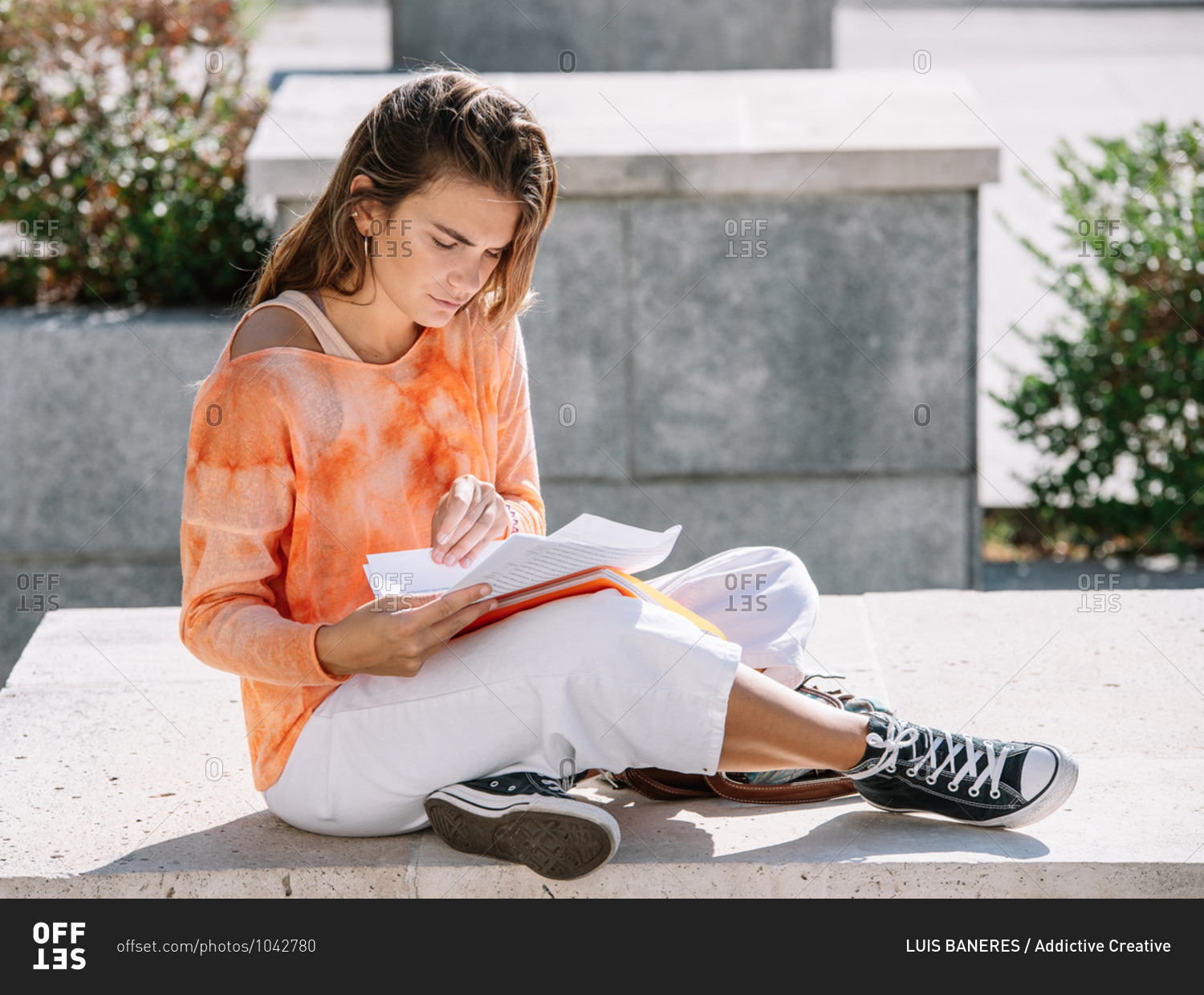 Low angle of young woman in casual clothes sitting cross legged on border and opening notepad while doing homework outside university building in Madrid, Spain