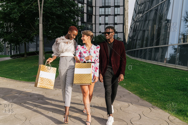 Full length of group of modern millennial multiethnic friends in fashionable outfits holding shopping bags while standing together against contemporary city buildings with glass walls