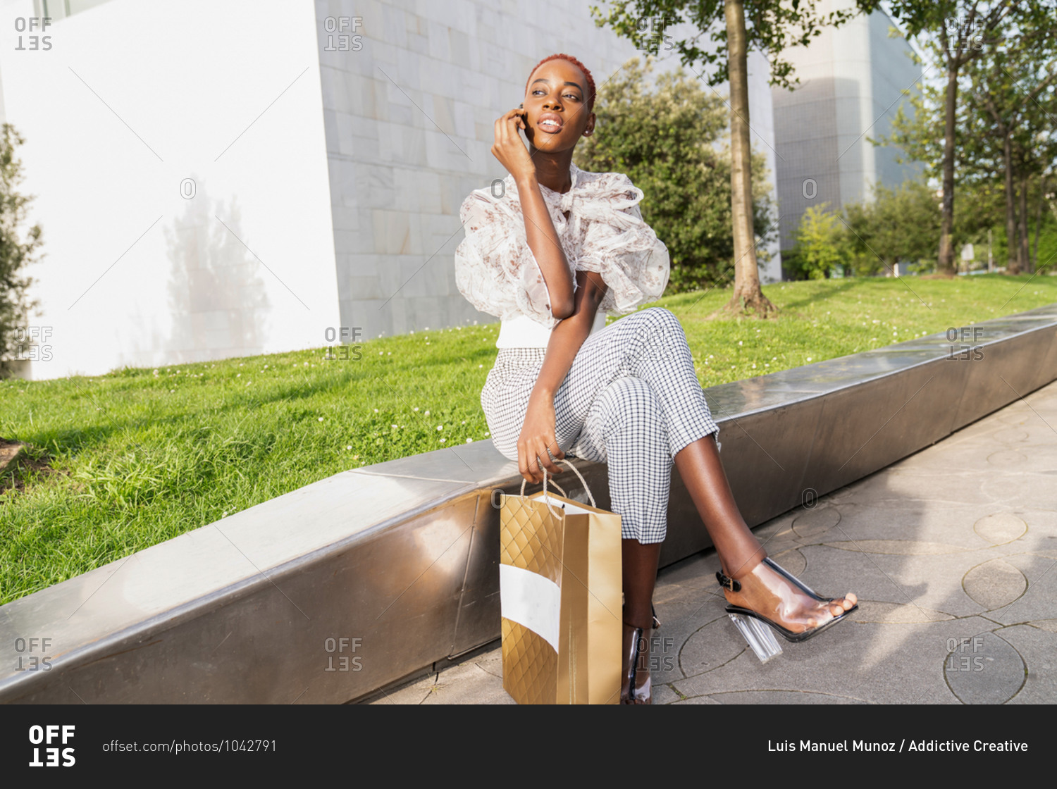 Young African American female with stylish haircut dressed in fashionable clothes and high heels with shopping bag in hand relaxing on stone border against modern urban building after shopping