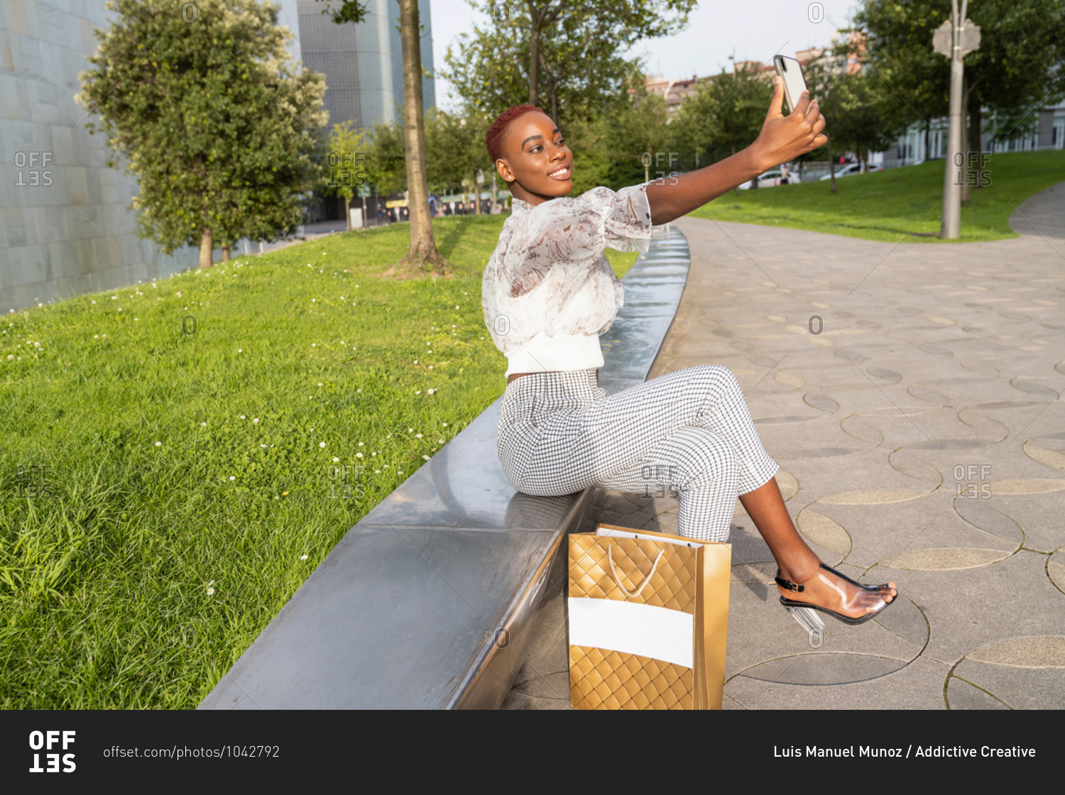 Young African American female with stylish haircut dressed in fashionable clothes and high heels with shopping bag taking a selfie relaxing with her mobile on stone border against modern urban building after shopping