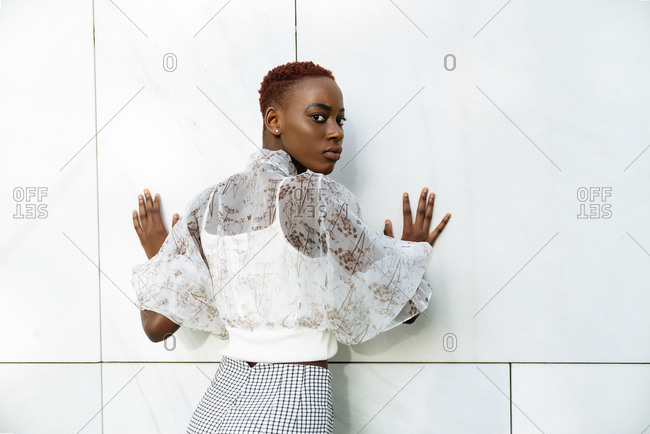 Crop of back view of confident young slim African American female in stylish blouse and pants standing against wall of modern building and looking at camera