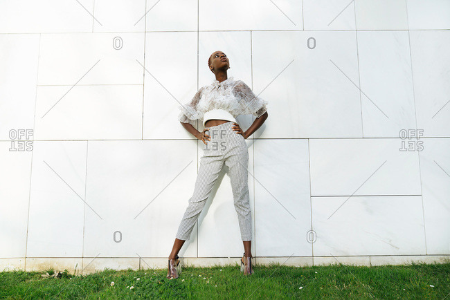 Low angle full body of confident young slim African American female in stylish blouse and pants standing against wall of modern building and looking up