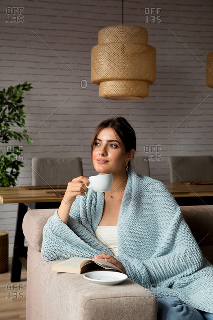 Pensive young female wrapped in warm blanket sitting on couch and enjoying hot tea while chilling in cozy living room at home