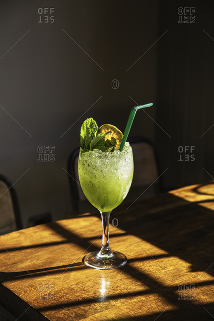 Glass of fresh appetizing icy cocktail with mint and lemon slice served with straw on wooden table in pub