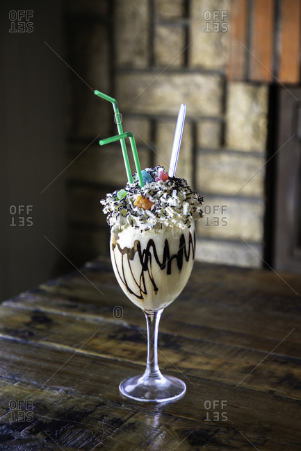Glass of delicious sweet milkshake with chocolate syrup and whipped cream garnished with colorful sprinkles served with straws on wooden table