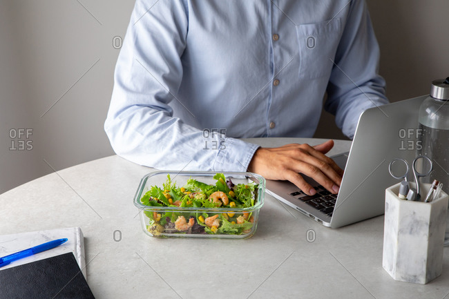 Crop view of unrecognizable office employee sitting at table in workplace and eating delicious salad with greenery and shrimps while having nutritious lunch
