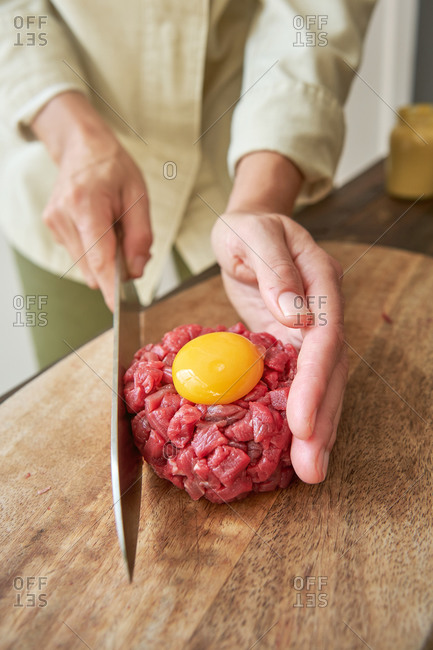 From above of unrecognizable female chef cooking Steak tartare in kitchen while making round shape of raw beef with egg yolk