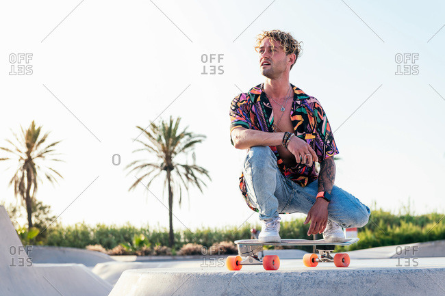 Low angle of young stylish male skater in colorful shirt and jeans sitting on skateboard while spending summer day in skatepark