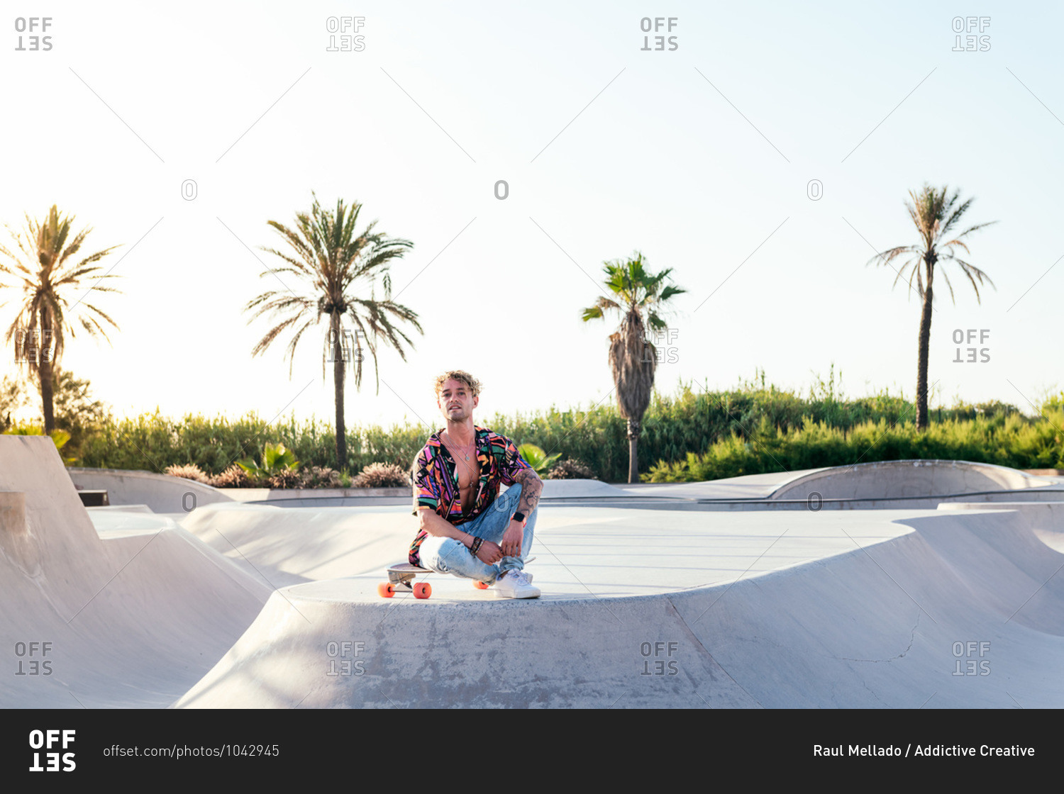 Low angle of young stylish male skater in colorful shirt and jeans sitting on skateboard while spending summer day in skatepark