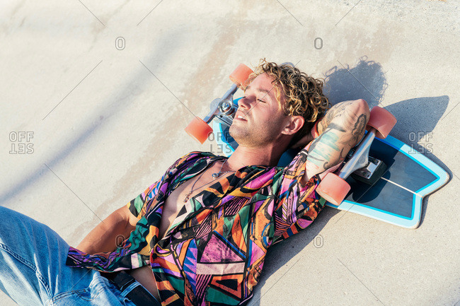 High angle of young hipster tattooed male in stylish colorful shirt lying on concrete ramp with skateboard under head and enjoying summertime in skatepark