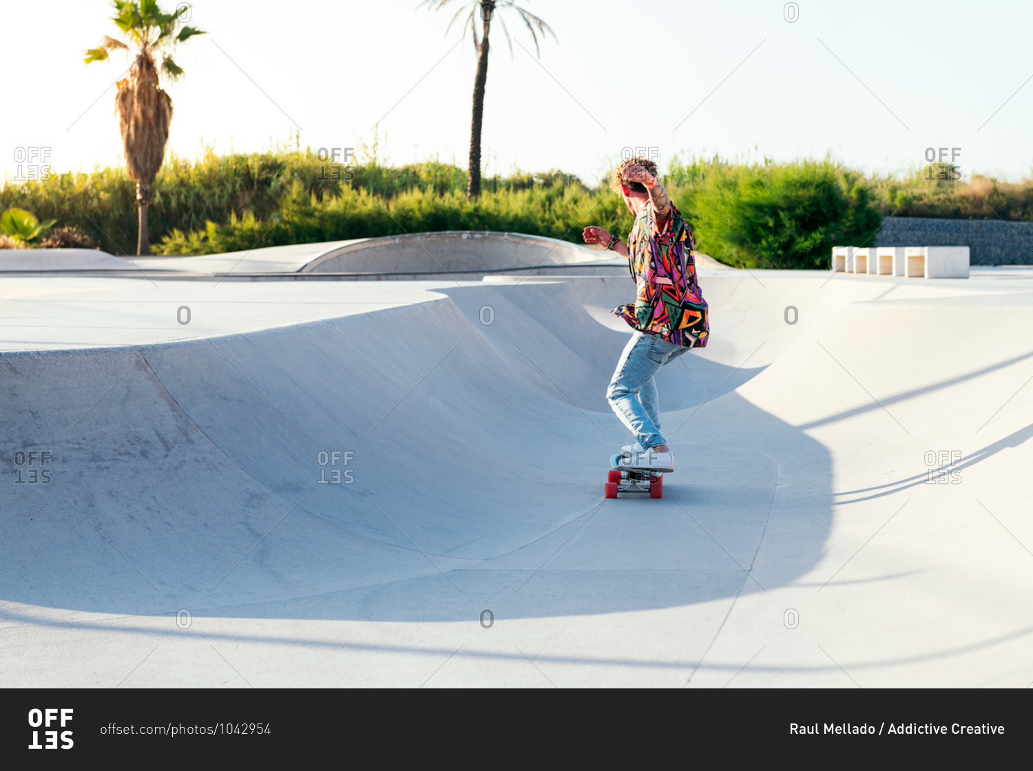 Back view of anonymous funky young male skateboarder in trendy colorful shirt and jeans performing trick on concrete ramp while practicing skills in skatepark