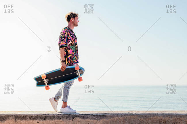 Low angle side view of young hipster male skater in trendy summer outfit carrying skateboard while walking on embankment against sea and cloudless sky in summer day
