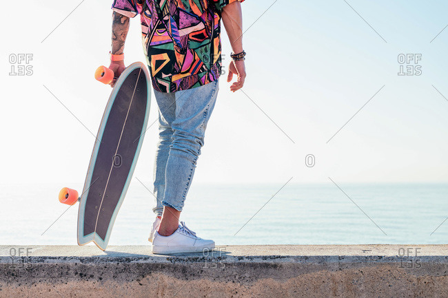 Unrecognizable stylish skater in jeans and trendy shirt standing holding a skateboard on street on sunny day in summer