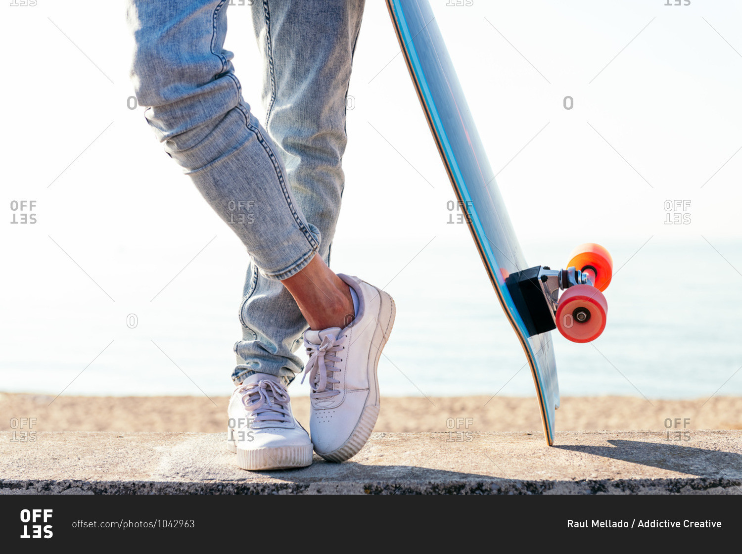 Unrecognizable stylish skater in jeans and sneakers standing next to skateboard on street on sunny day in summer