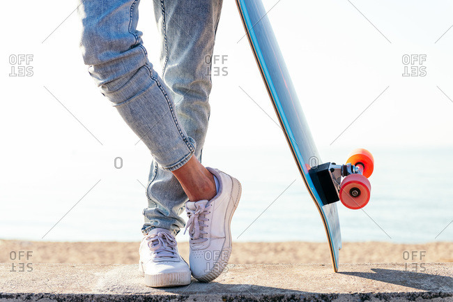 Unrecognizable stylish skater in jeans and sneakers standing next to skateboard on street on sunny day in summer