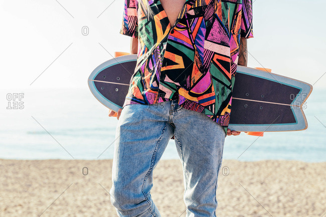 Crop view of anonymous young hipster male skater in trendy summer outfit carrying skateboard while standing on embankment against sea and cloudless sky in summer day