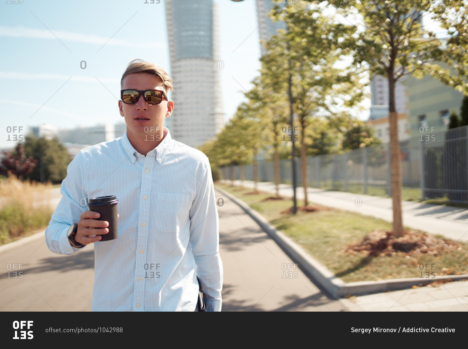 Confident male entrepreneur in stylish sunglasses walking along street with coffee to go during break in city