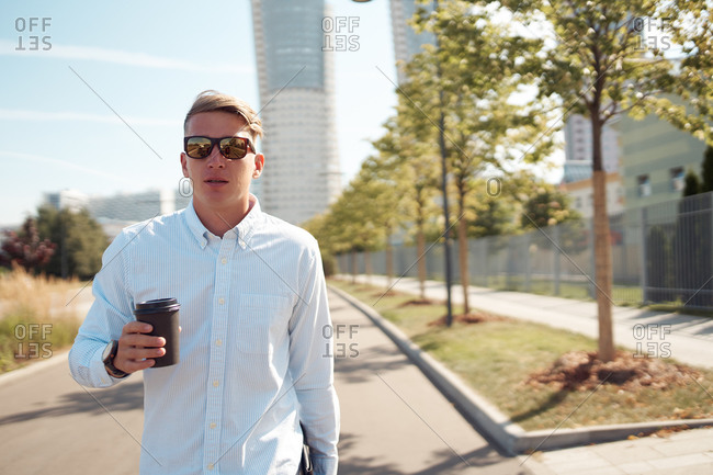 Confident male entrepreneur in stylish sunglasses walking along street with coffee to go during break in city