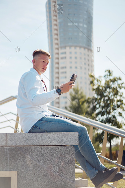 Low angle side view of carefree male in stylish wear sitting on stone border in city and reading messages on cellphone while enjoying weekend
