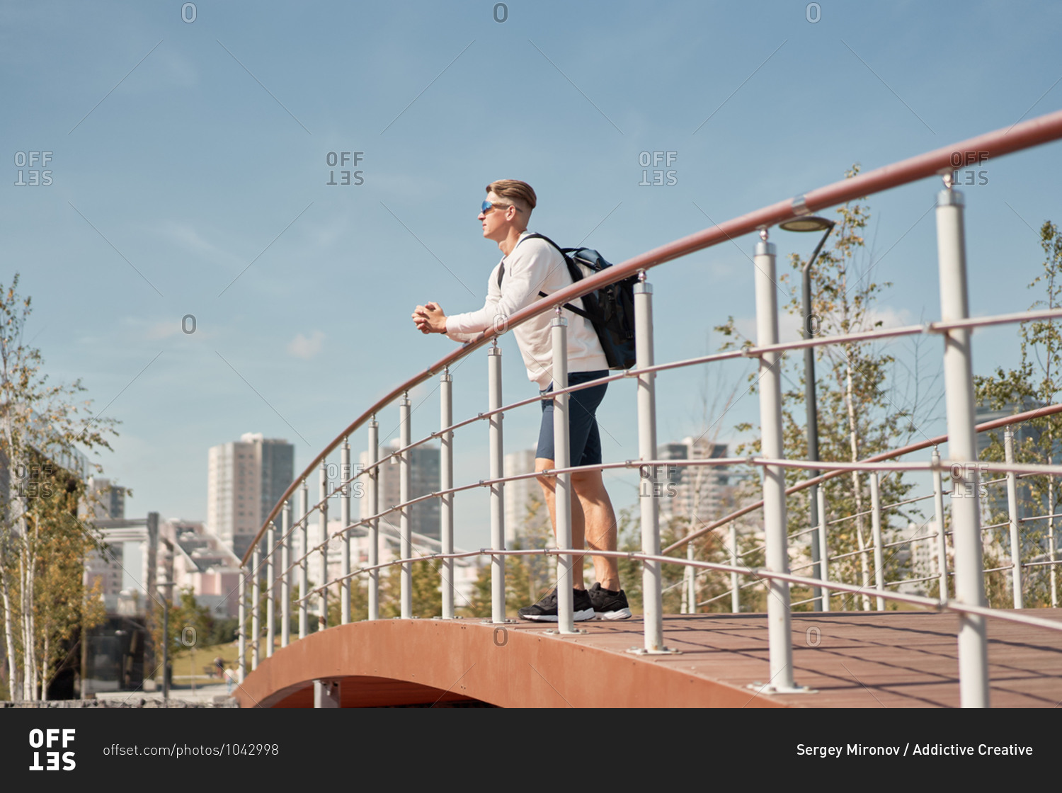Side view of young male tourist in casual clothes and sunglasses with backpack leaning on railing of pedestrian bridge and looking away while resting after walking in city in summer day