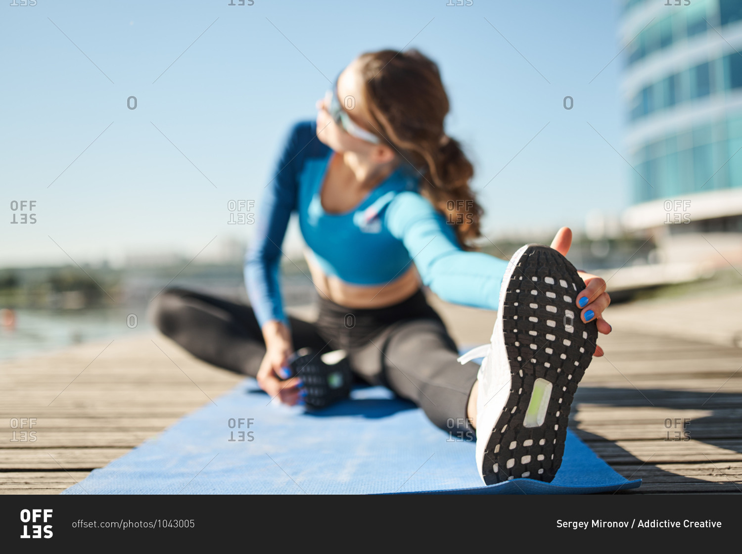 Unrecognizable slim female in activewear and sport shoes sitting on mat and stretching legs during outdoor fitness workout on urban embankment