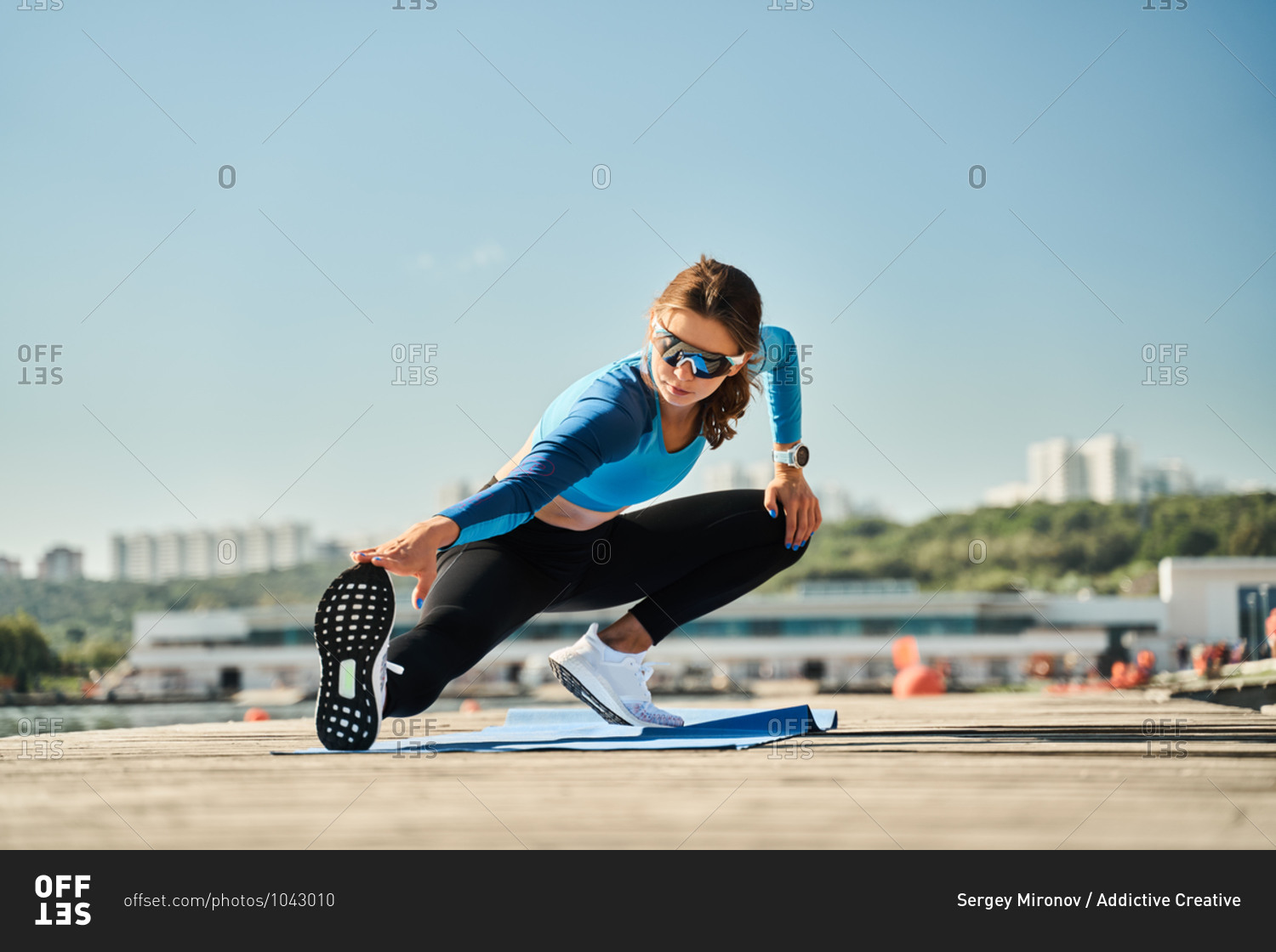 From above active slim female in sportive outfit standing on mat and doing stretching exercise for legs while training on wooden pier near river