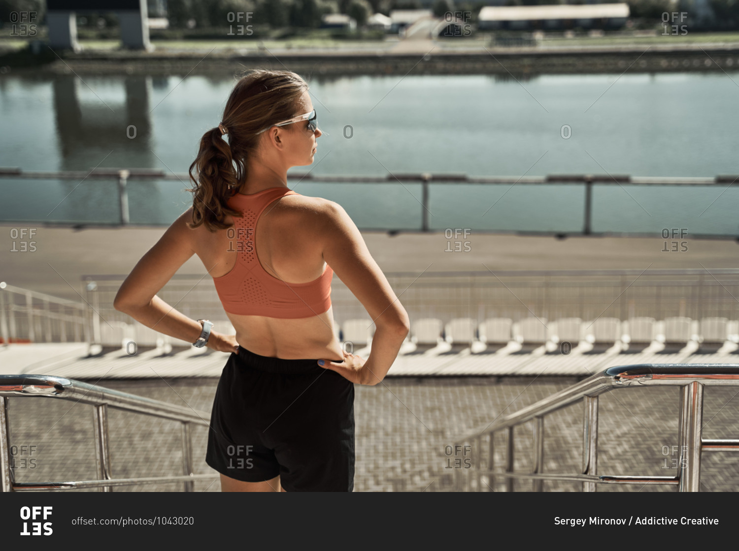 Back view of unrecognizable slim female athlete in sportswear standing on urban embankment near river and relaxing after exercising in summer day