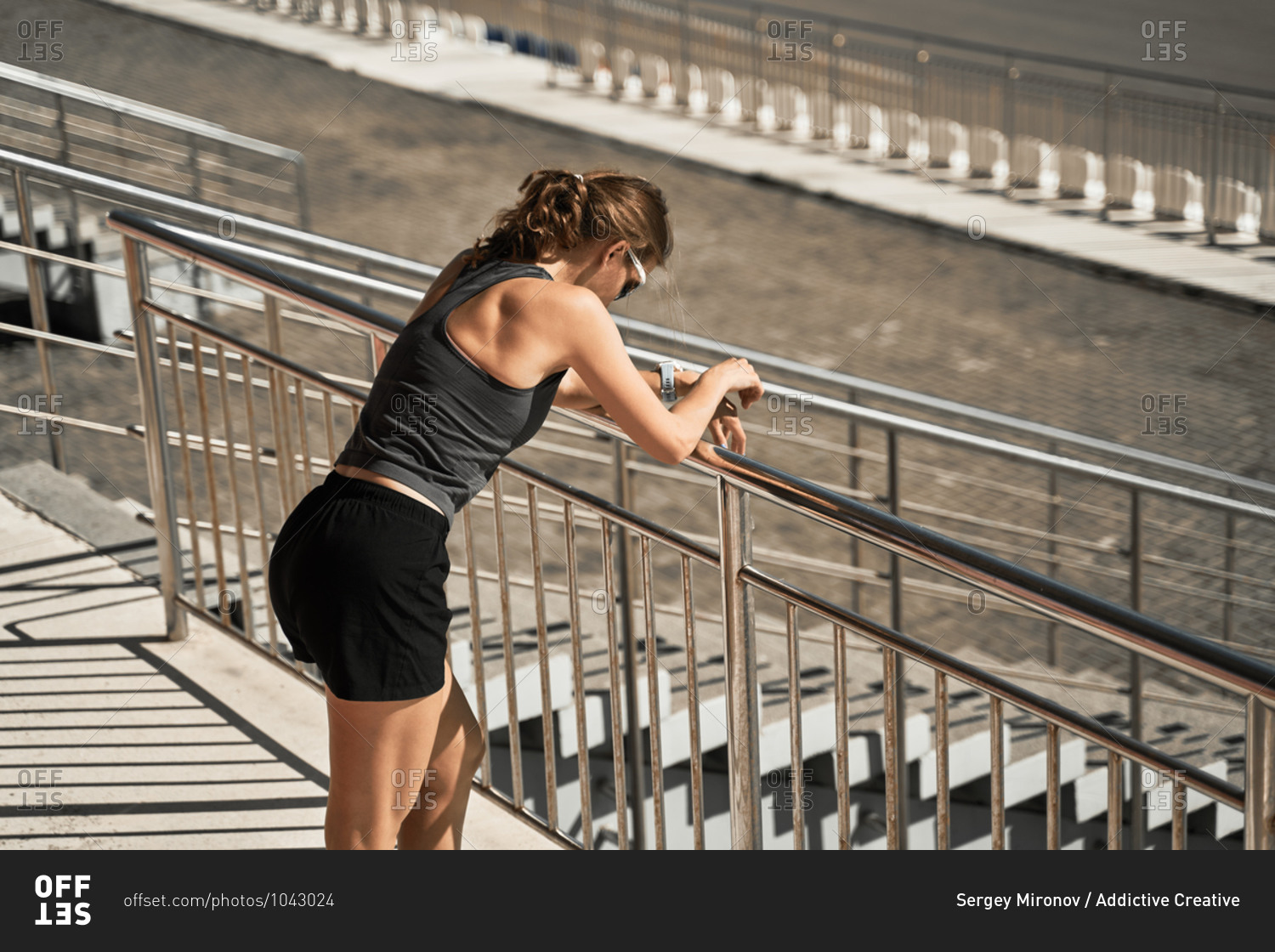 Side view of tired young sportswoman in black sportive outfit and sunglasses leaning forward on railing on stadium tribune while resting after outdoor workout
