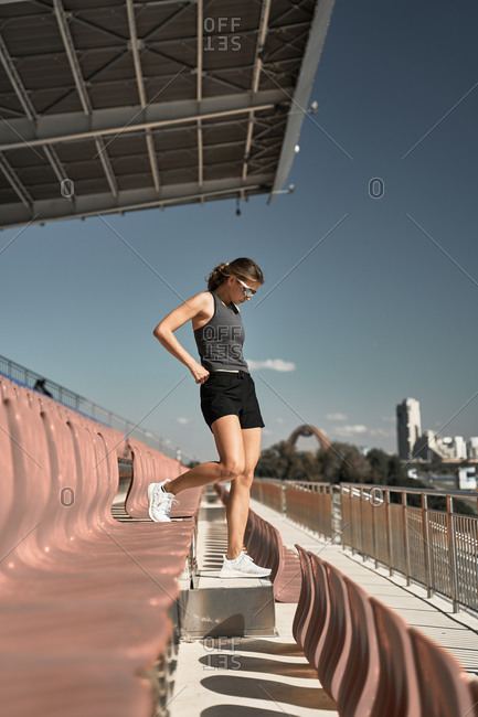 Full body side view of determined young sportswoman in activewear and sunglasses walking along tribune of stadium against blue sky