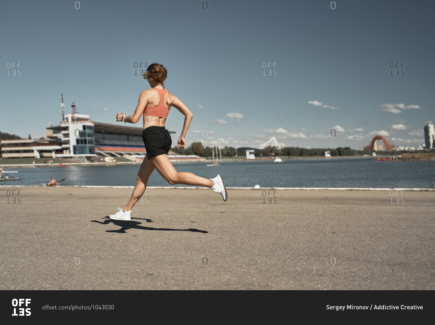 Side view full body of energetic motivated fit female in activewear with fitness tracker jogging on paved embankment near river in city