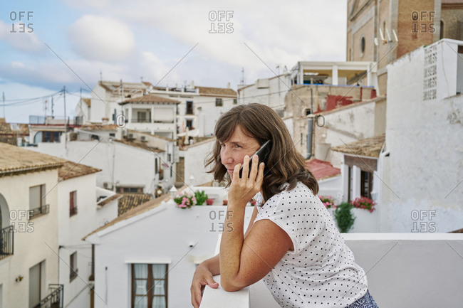Side view of mature female standing on balcony and talking on smartphone on background of cityscape of Altea while looking away