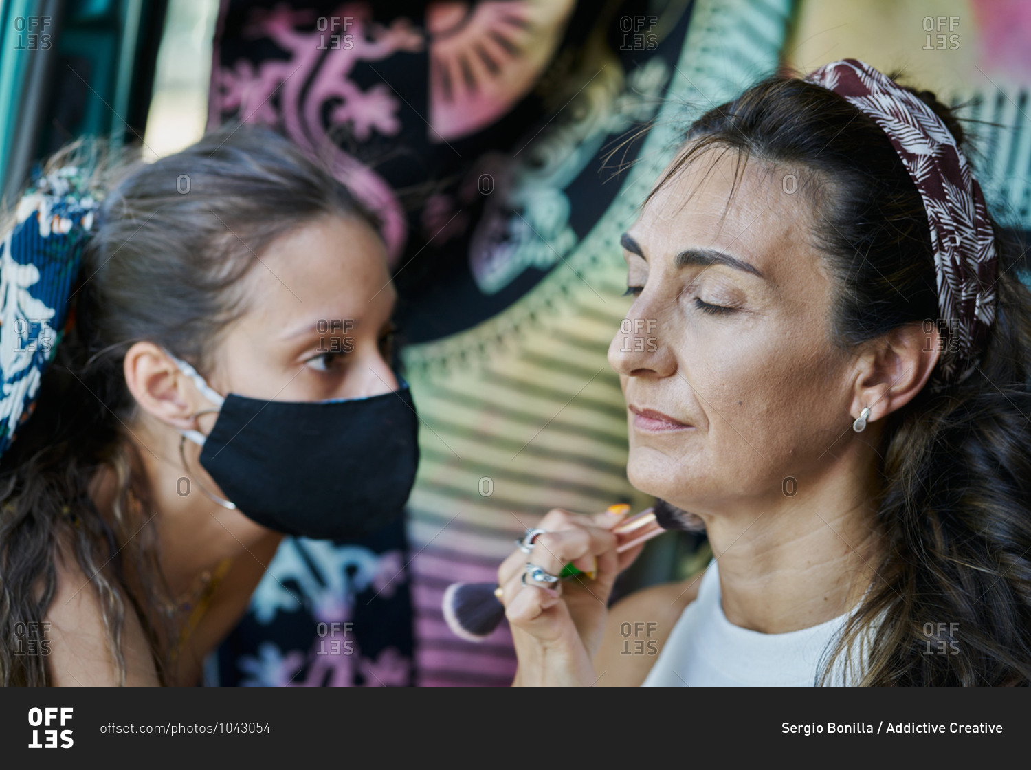 Female artist with face mask applying foundation with brush on face of mature female while doing stylish makeup